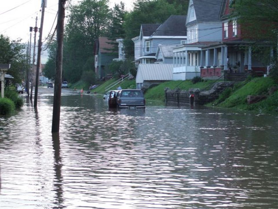 Flooded street with a dark SUV partially submerged in water. Houses line the street