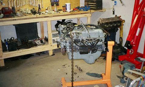1989 Ford Bronco - An engine is suspended on an orange engine stand in a cluttered workshop, with tools on a workbench in the background.