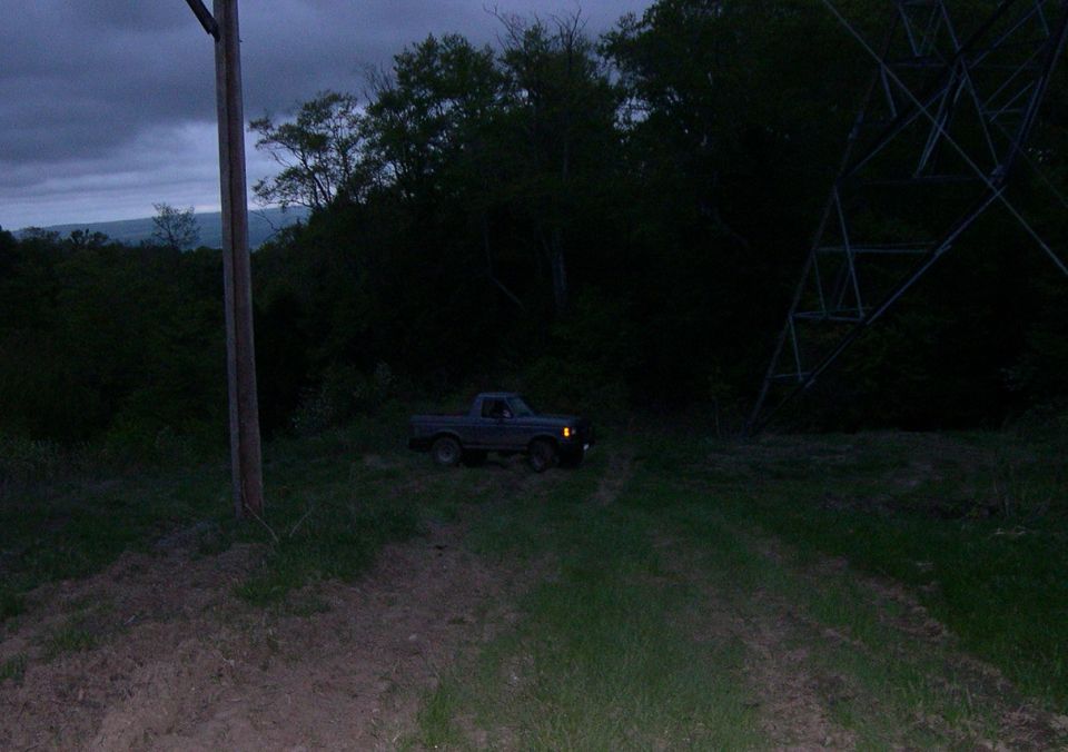 1989 Ford Bronco parked on a dirt path next to a power line tower at dusk