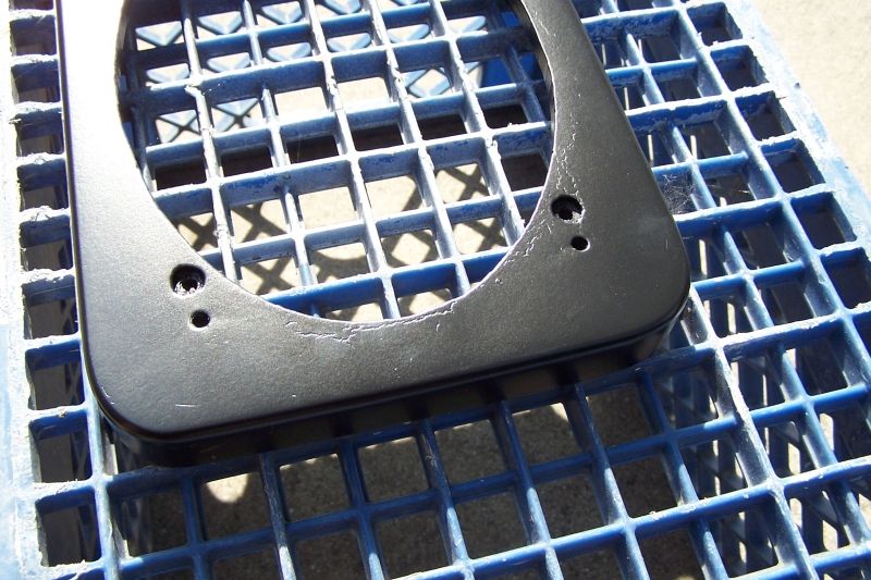 Black metal speaker adapter bracket with two mounting holes, resting on a blue plastic grid