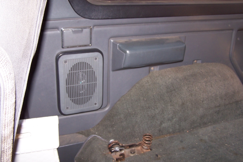 Interior view of a gray plastic panel with a speaker grille and a gray armrest above worn gray carpet