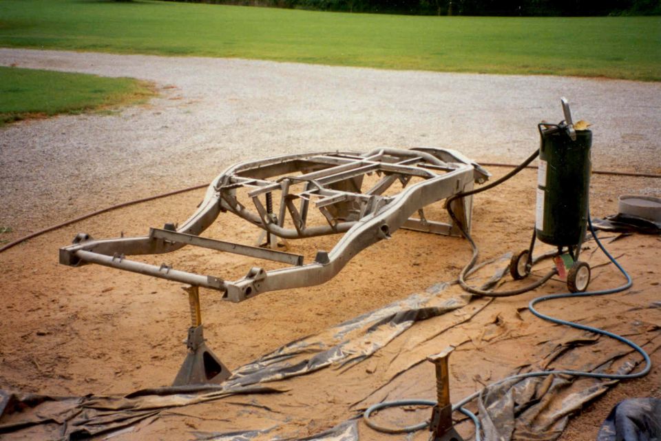 Blasted bare sports car frame on jack stands beside sandblasting pot outdoors