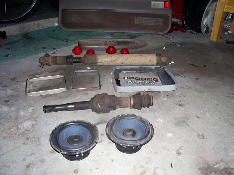 1991 Ford Bronco - Various automotive parts, including driveshafts and speakers, are displayed on a concrete floor next to a car door panel.