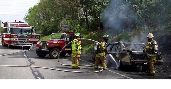 Firefighters are putting out a car fire on the side of the road. A fire truck and a Jeep are also present