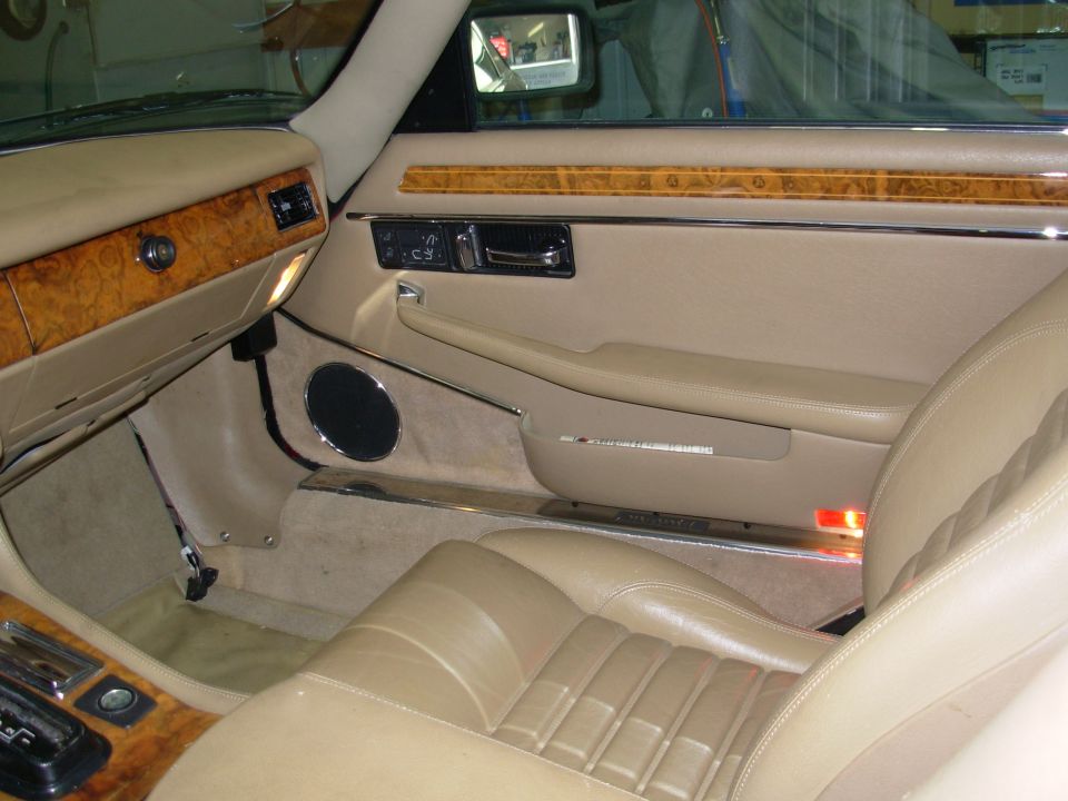 Tan leather interior of a Jaguar XJ-S showing passenger door with wood trim, chrome accents, and pleated seat