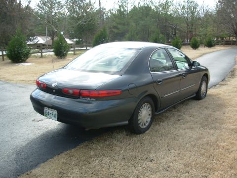 1999 Mercury Sable - A dark sedan is viewed from its rear-left quarter, parked on a driveway with dry grass and trees in the background, trees and a fence.