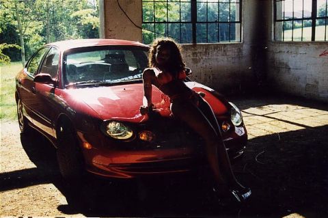 1998 Ford Taurus - A woman poses on the hood of a red sedan inside a garage with sunlight streaming through the windows.