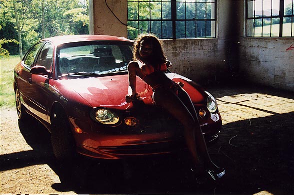 Woman on hood of red sedan. The car has a rounded front end and oval headlights
