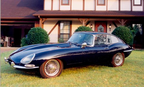 1967 Jaguar XKE - A 1967 Jaguar XKE coupe, finished in dark blue and equipped with wire wheels, is parked on a grassy lawn in front of a Tudor-style house.