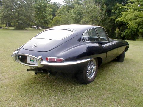 1967 Jaguar XKE - A dark blue 1967 Jaguar XKE coupe is viewed from the rear-right angle, parked on a grassy lawn with trees in the background.