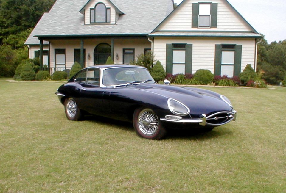 Dark blue Jaguar E-Type coupe with chrome wire wheels parked on a lawn in front of a house