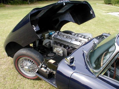 1967 Jaguar XKE - The engine bay of a dark blue Jaguar E-Type is visible with the hood open, revealing the inline-six engine and wire wheels.