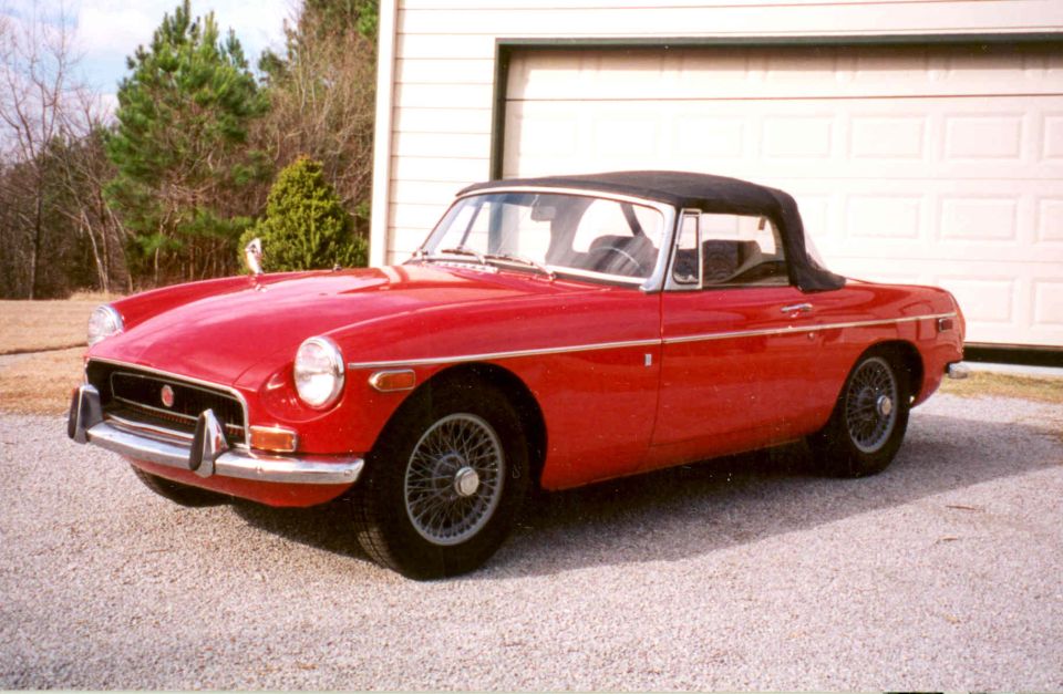Red convertible roadster with black top up and chrome wire wheels on a gravel driveway