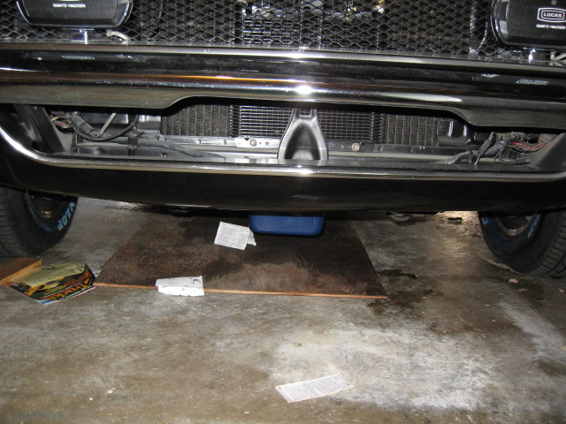 Car's front bumper, grille, and radiator are visible. An oil pan sits under the engine, with some papers and a blue container nearby