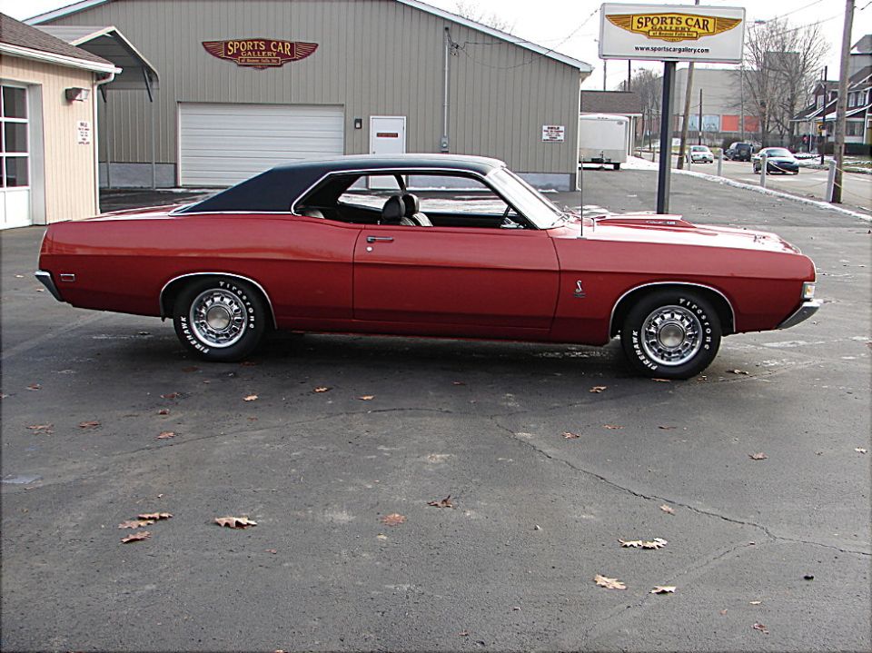 Red Ford Torino Cobra fastback with black vinyl roof, side profile outside a classic car dealership