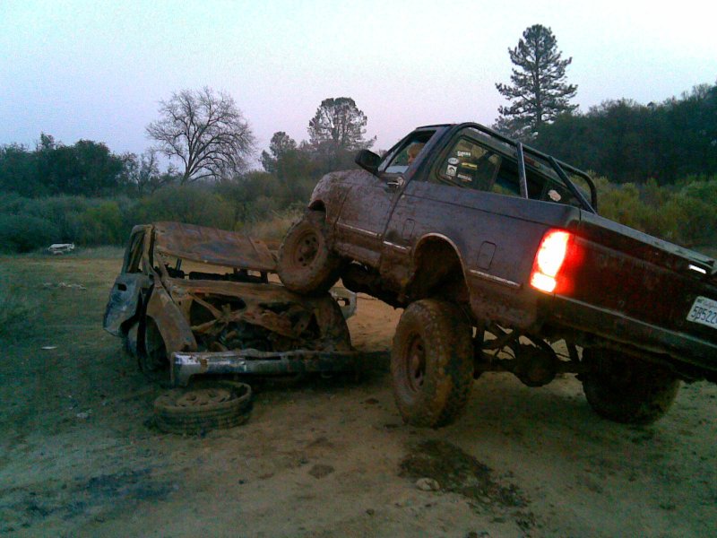 Muddy pickup truck with roll cage on top of a wrecked car. License plate 5B522Z