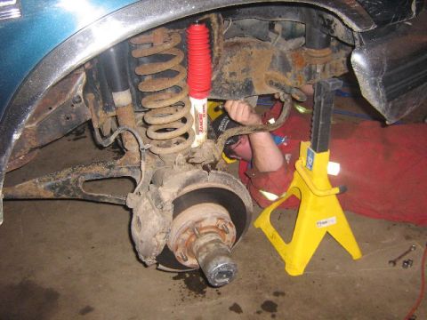 1995 Ford Bronco - A mechanic works on the suspension of a vehicle, which is supported by a yellow jack stand. A red Rancho shock absorber is visible.