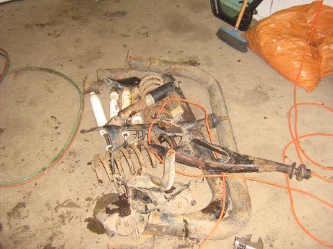 1995 Ford Bronco - Various rusty vehicle suspension parts are scattered on a garage floor next to an orange bag and a broom.