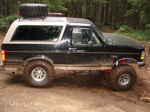 1996 Ford Bronco - A black and tan Ford Bronco with a spare tire on the roof is driven off-road on a muddy trail.