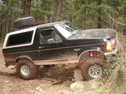 1996 Ford Bronco - A lifted Ford Bronco navigates a muddy, wooded trail with its tires kicking up dirt.