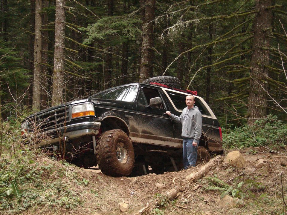 Black Ford Bronco with lifted suspension and large tires on a dirt trail, a person stands beside the driver's door