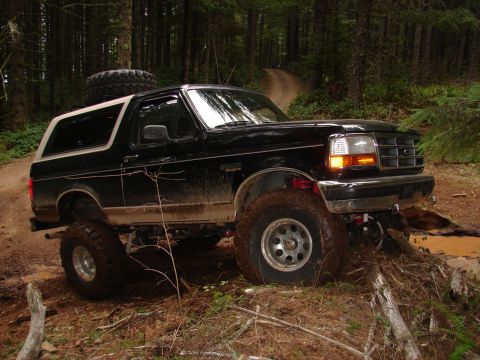 1996 Ford Bronco - A black 1996 Ford Bronco navigates a muddy, wooded trail with a spare tire mounted on the roof.