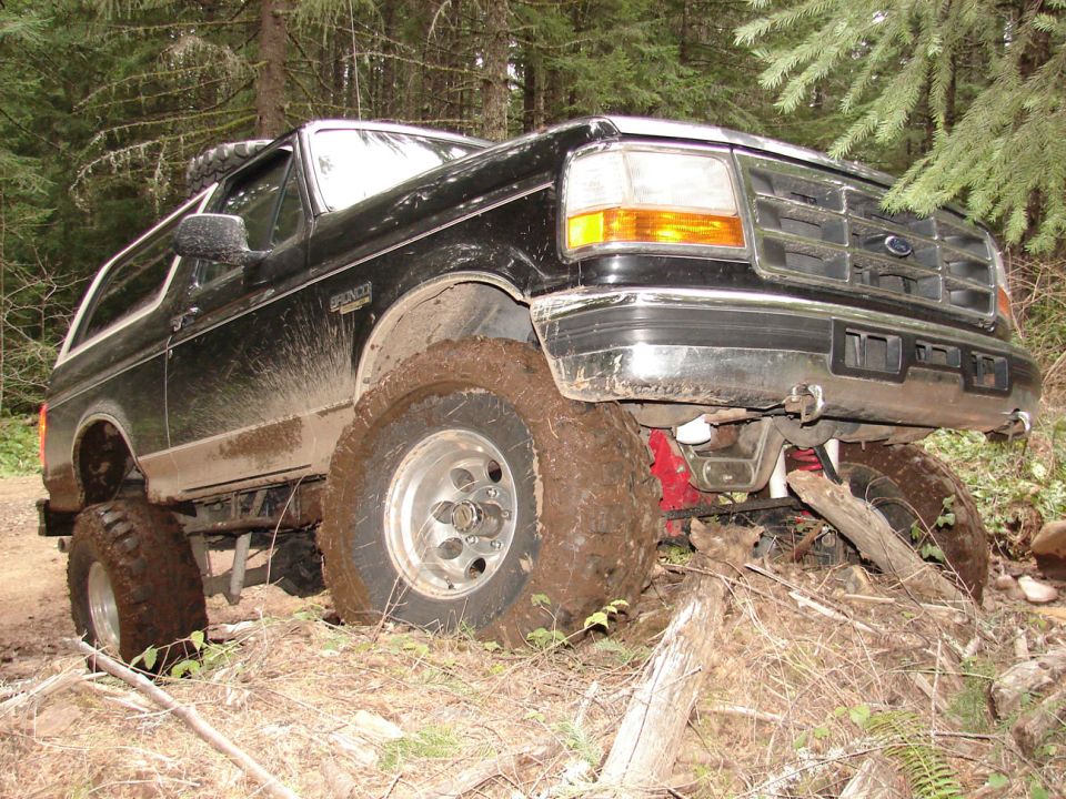 Black 1996 Ford Bronco with large tires stuck in mud on a wooded trail