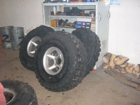 1995 Ford Bronco - A collection of off-road tires and rims are stored indoors, with a shelf unit and firewood nearby.
