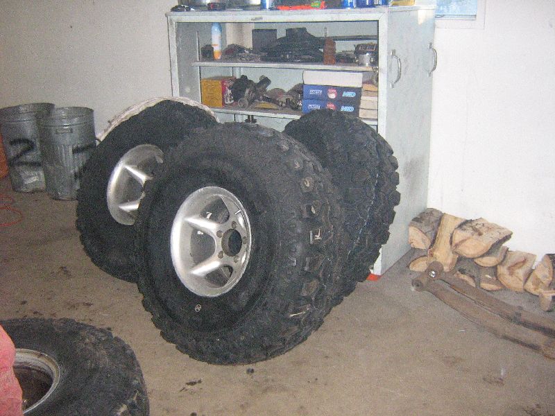 Several off-road tires stacked up, with one in the foreground. A metal shelf unit is in the background