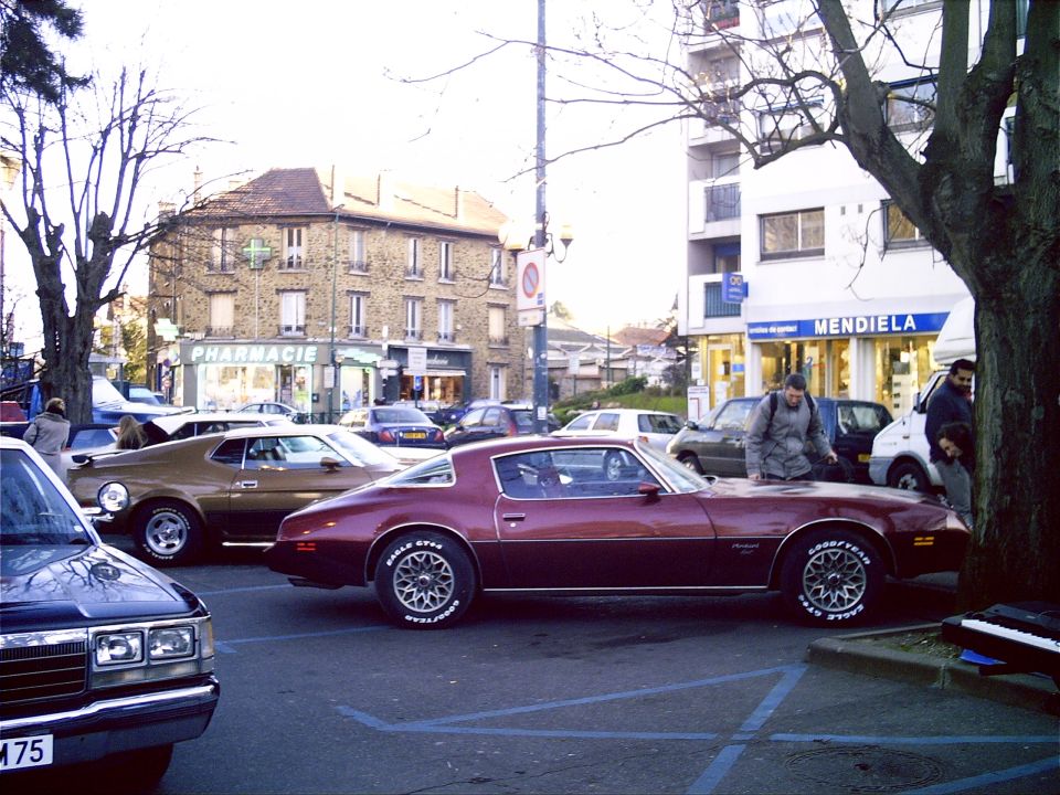 Maroon Pontiac Firebird and brown coupe parked in a European town square