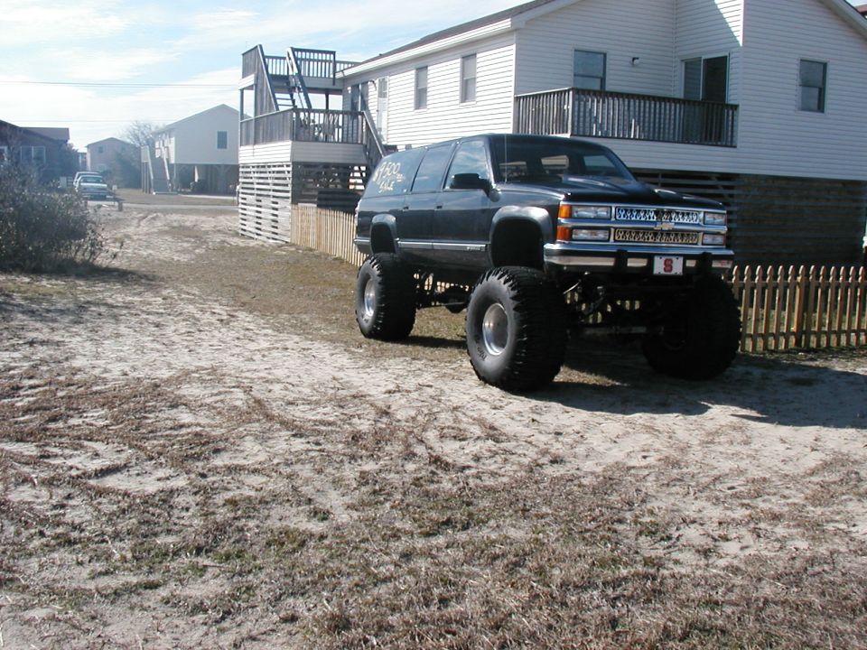 Black lifted Chevrolet Suburban on large tires parked on sandy ground by houses