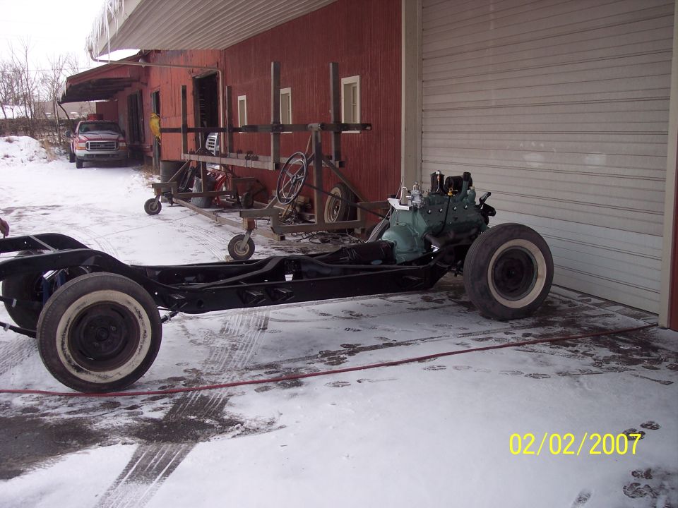 Black restored rolling chassis with green engine on snowy pavement beside a red barn
