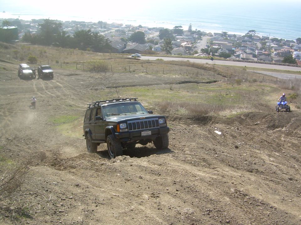 Dark green Jeep Cherokee XJ with roof rack driving off-road, front grille visible