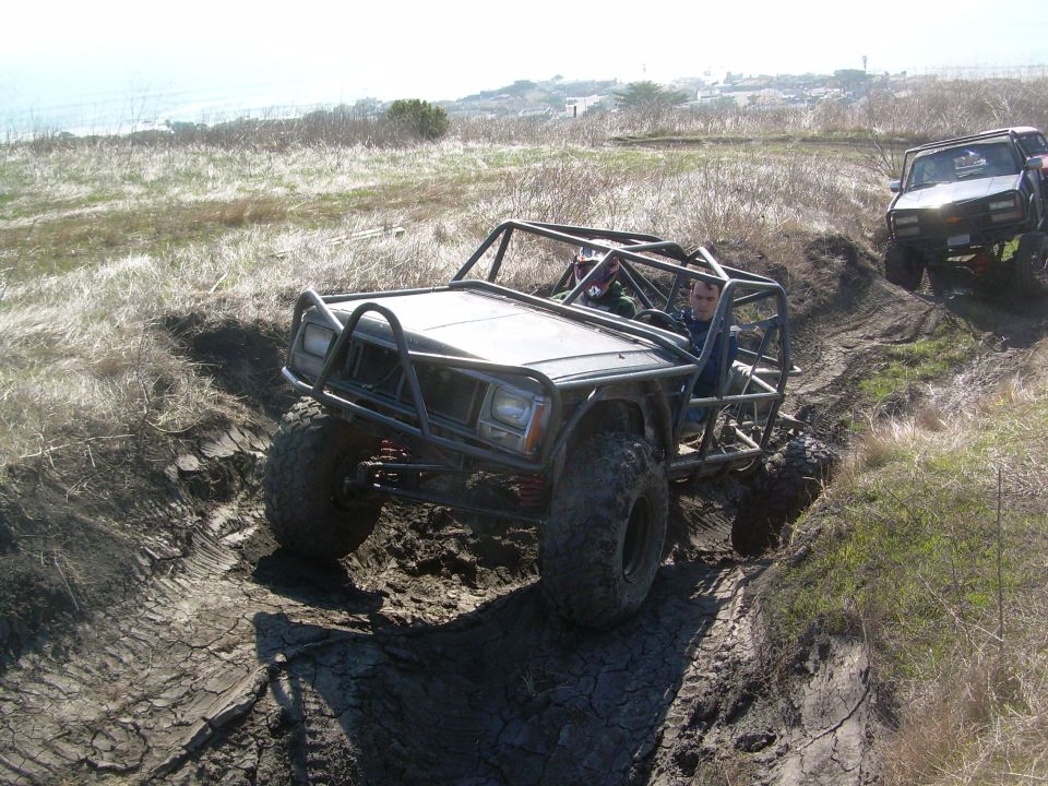 Off-road buggy with a roll cage climbing a muddy hill. Another vehicle is behind it