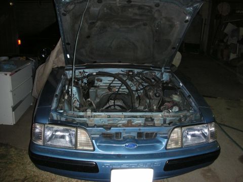 1990 Ford Mustang - The engine bay of a blue Ford Mustang is shown with the hood open, revealing the engine and various components.