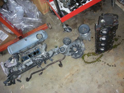 1990 Ford Mustang - Disassembled engine components, including a block and intake manifold, lie on a concrete floor next to a red engine stand.