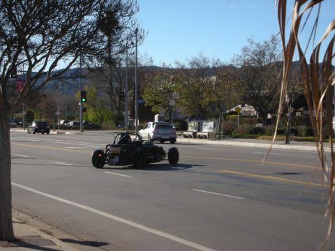 1998 Ford Crown Victoria - black skeletal open-wheel kit car drives down a sunny multi-lane boulevard with bare trees and hills behind it.
