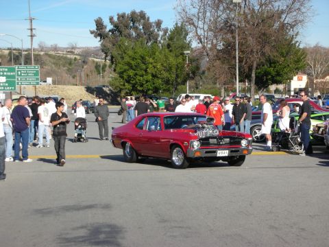 1998 Ford Crown Victoria - maroon classic Chevrolet Nova with a chrome supercharger protruding through the hood rolls through a crowded car-show parking lot.