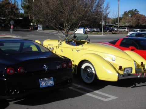 1998 Ford Crown Victoria - yellow speedster-style boattail roadster with whitewall wire wheels parked between a black Ferrari and a red car at a meet.