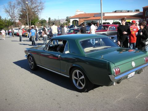 1998 Ford Crown Victoria - green early Ford Mustang coupe shown in rear three-quarter view among spectators and parked cars at an outdoor meet.