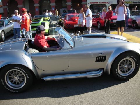 1998 Ford Crown Victoria - silver Shelby Cobra-style roadster with side pipes and a driver seated, parked in front of Hooper Camera at a street car show.