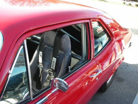 1998 Ford Crown Victoria - View through a red muscle car's side window revealing a roll cage and black racing bucket seat with harness.