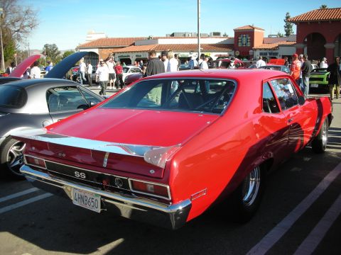 1998 Ford Crown Victoria - red Chevrolet Nova SS with black side stripe shown rear three-quarter in a busy car-show lot with Spanish-style buildings behind.