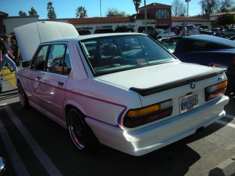 1998 Ford Crown Victoria - white BMW sedan with M tricolor stripe, rear spoiler, and personalized plate parked rear three-quarter at an outdoor car show.