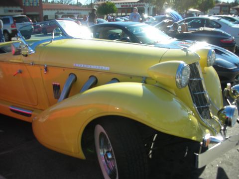 1998 Ford Crown Victoria - Close-up of a yellow supercharged boattail speedster's front fender, chrome grille, headlamps, and side exhaust pipes.