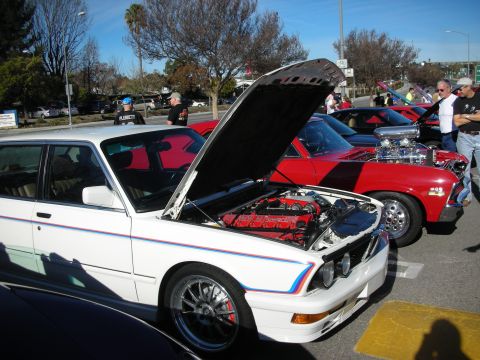 1998 Ford Crown Victoria - white BMW sedan sits with its hood open next to a red supercharged Nova among show cars and spectators in a sunny lot.