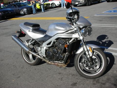 1998 Ford Crown Victoria - silver Triumph naked sportbike with a helmet resting on the seat stands in a sunlit parking lot.