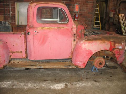 1952 Ford Pickup - Side view of a rusty red pickup truck body, missing its front wheel and hood, resting on jack stands.