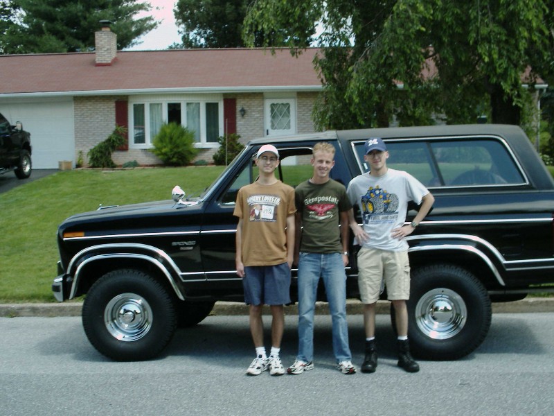 Three guys stand in front of a black Ford Bronco. A house and lawn are in the background