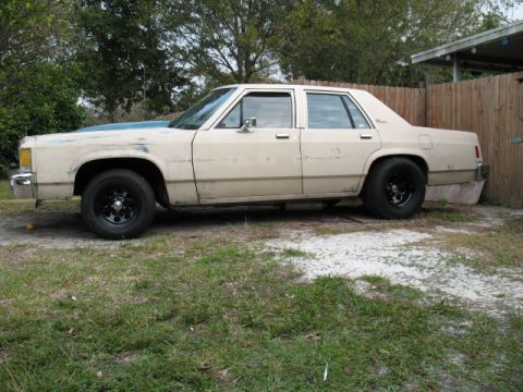 1985 Ford LTD Crown Victoria - This tan sedan, possibly a Ford LTD, sits parked outdoors with black aftermarket wheels and a contrasting blue hood.
